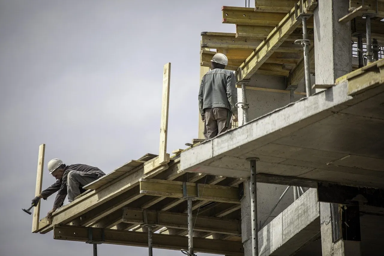 Zwei Bauarbeiter mit Helm arbeiten auf einem Betongerüst mit Holzschalungen an einem mehrgeschossigen Rohbau.