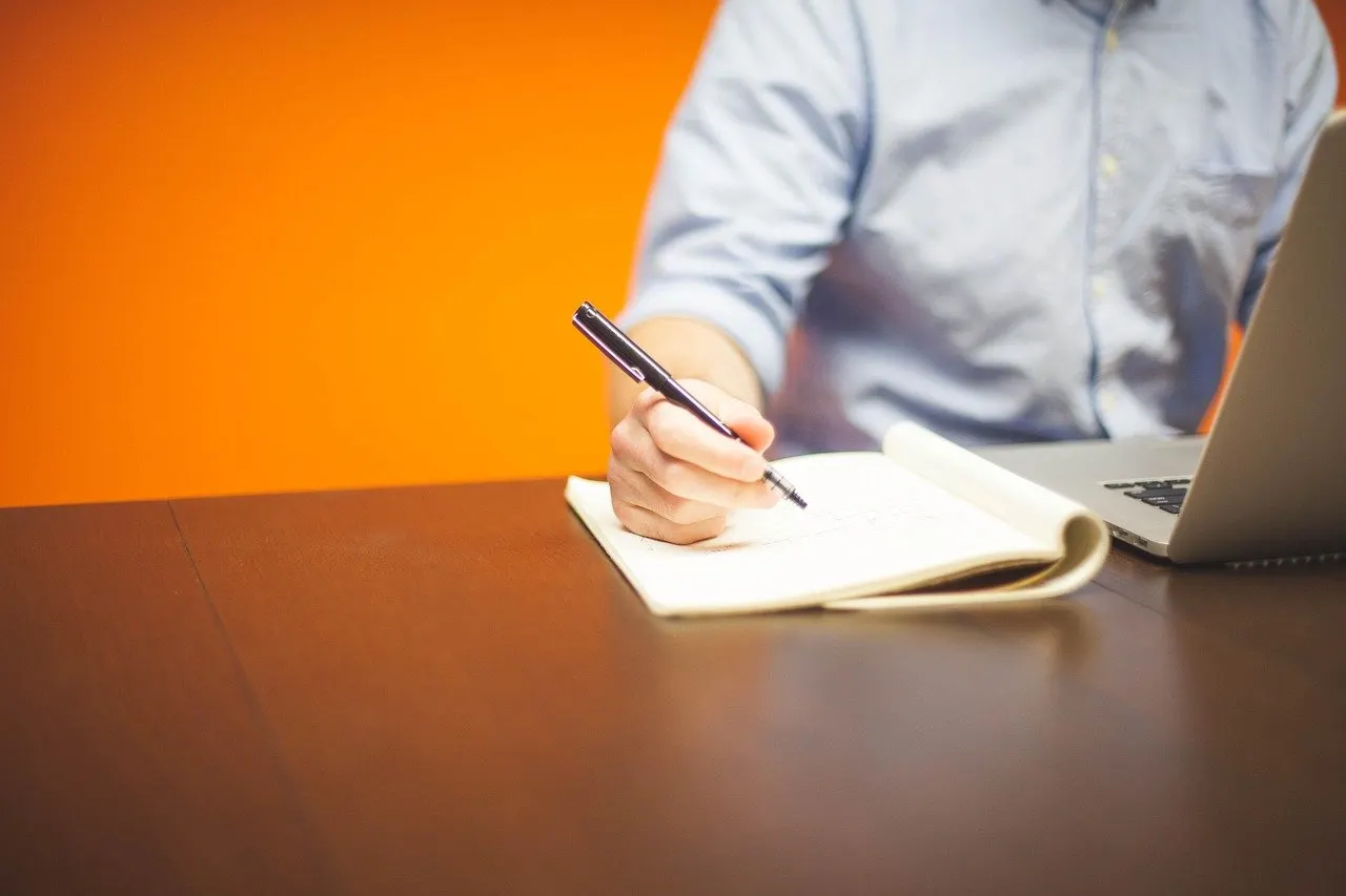 Person in hellblauem Hemd notiert mit Stift in ein Notizbuch, daneben Laptop, vor oranger Wand.