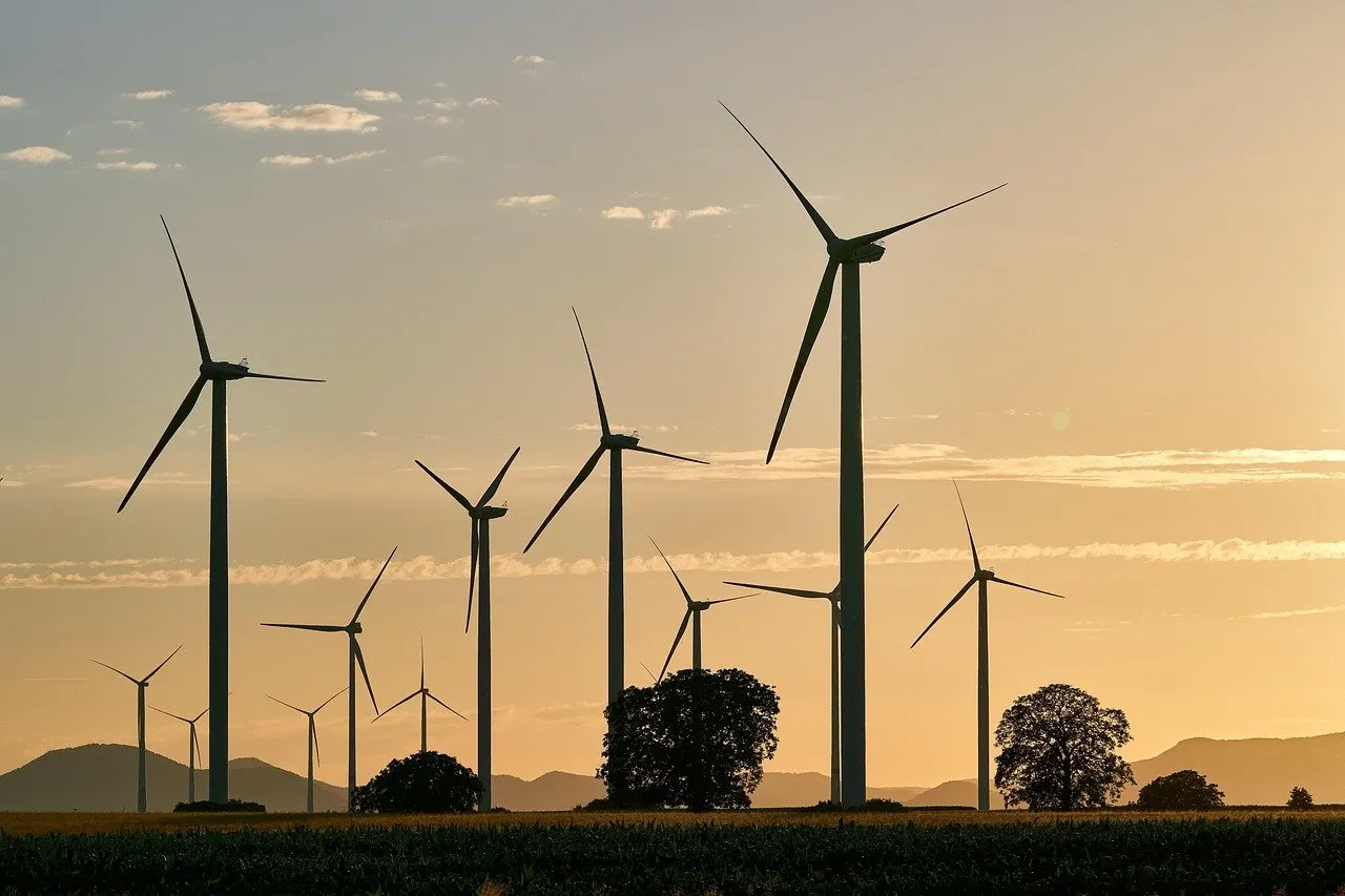 Mehrere Windkraftanlagen auf einem Feld vor einem orangefarbenen Abendhimmel, davor Bäume und Hügel.