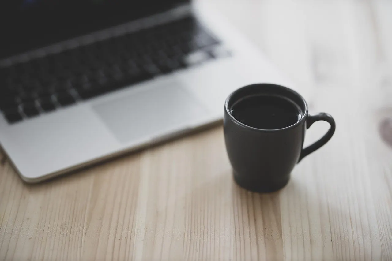 Schwarze Tasse neben aufgeklapptem Laptop auf einem hellen Holztisch.