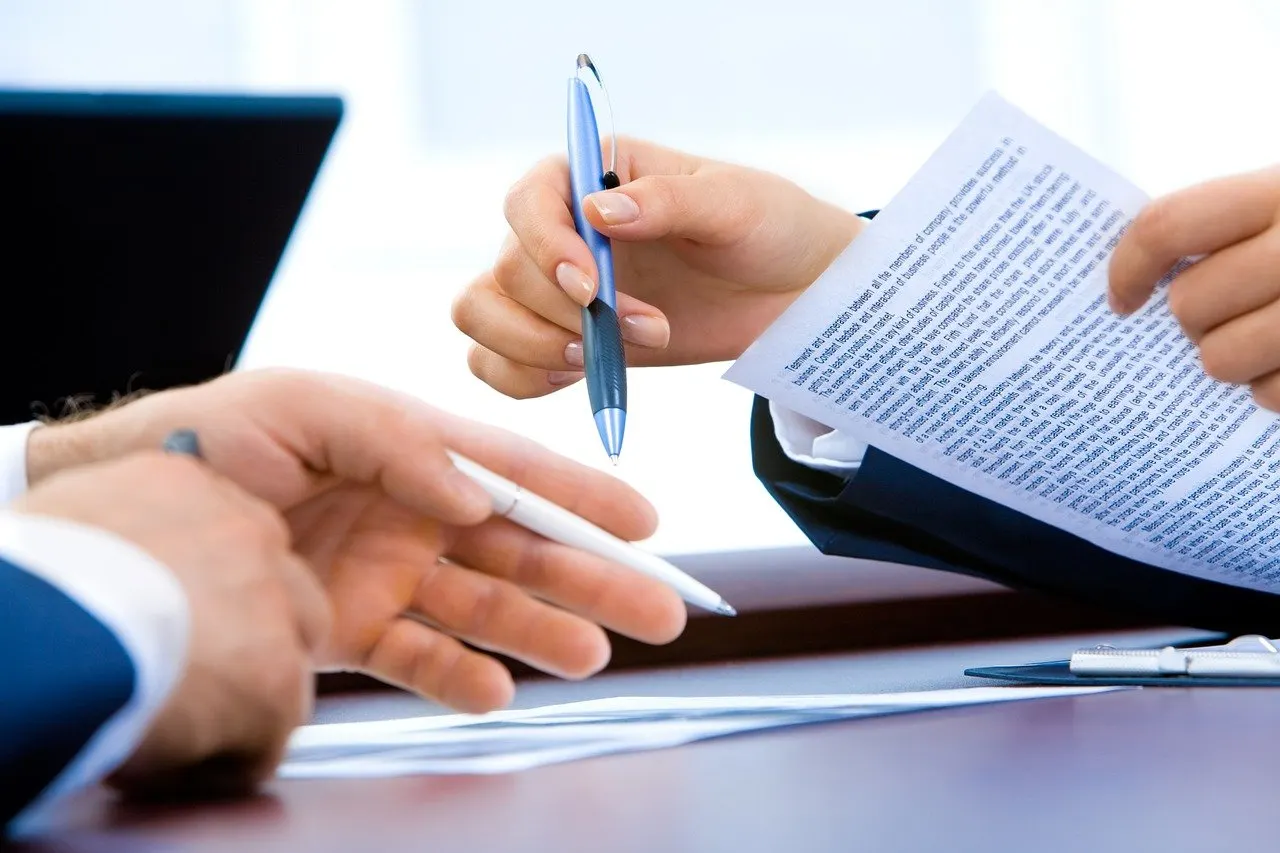 Zwei Personen am Tisch, eine reicht ein Blatt mit Text, ein Stift wird übergeben